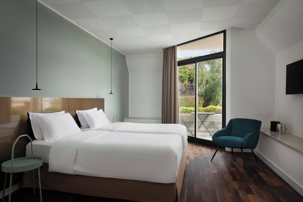 Hotelkamer met twee bedden, een televisie, een stoel en een balkon met uitzicht op groen.
