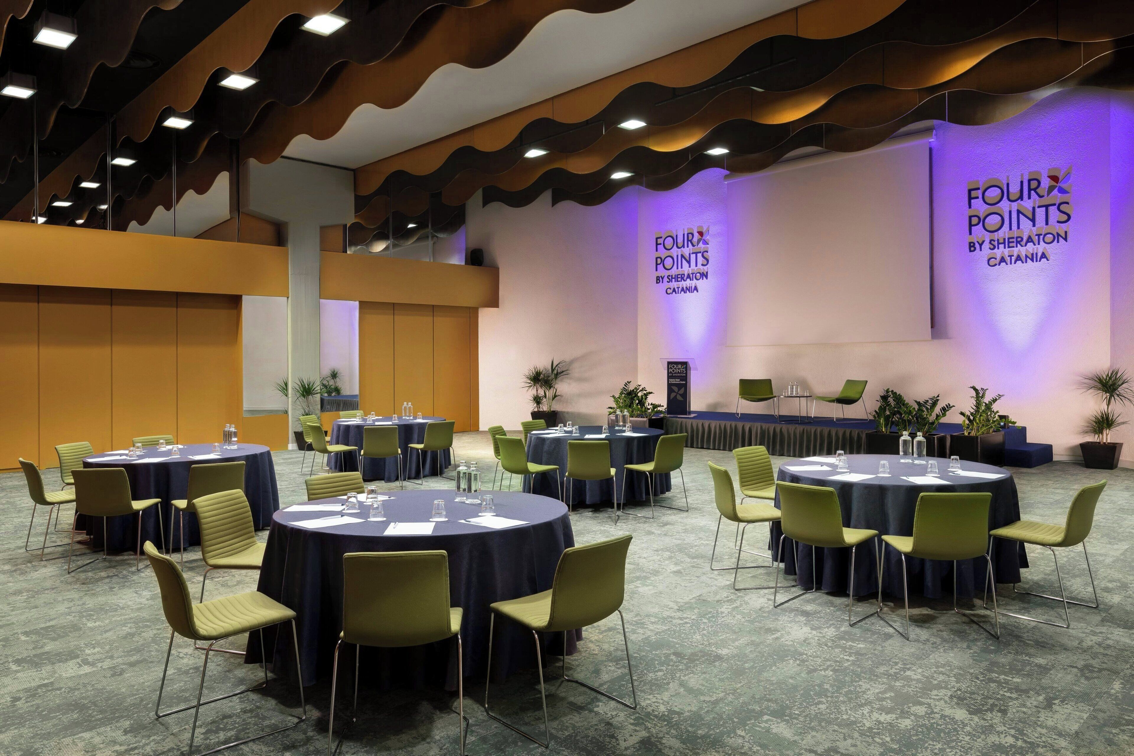Un salón de conferencias con mesas y sillas redondas, un escenario y una pared con el texto "FOUR POINTS BY SHERATON CATANIA".