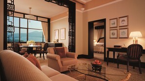 Quarto de hotel moderno com sofá, poltronas, mesa de centro de vidro e área de jantar com vista para as montanhas.