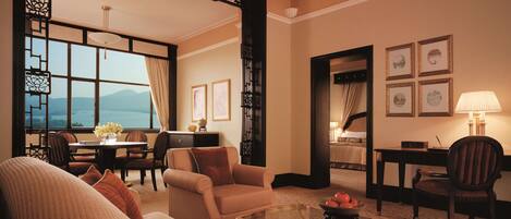 A modern hotel room with a sofa, armchairs, a glass coffee table, and a dining area with a view of the mountains.