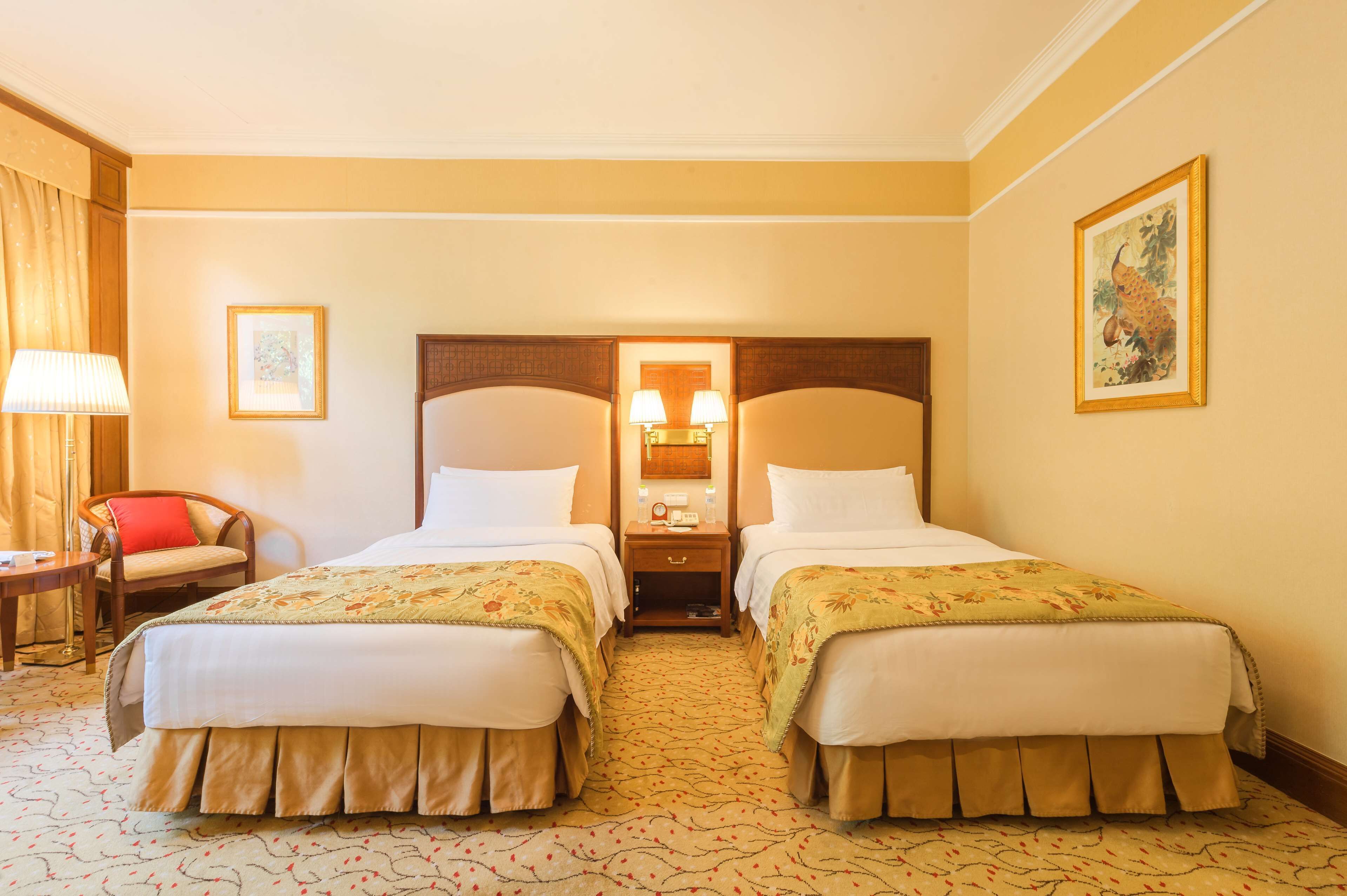 A hotel room with two beds, a nightstand with a lamp, a chair, and a painting on the wall.