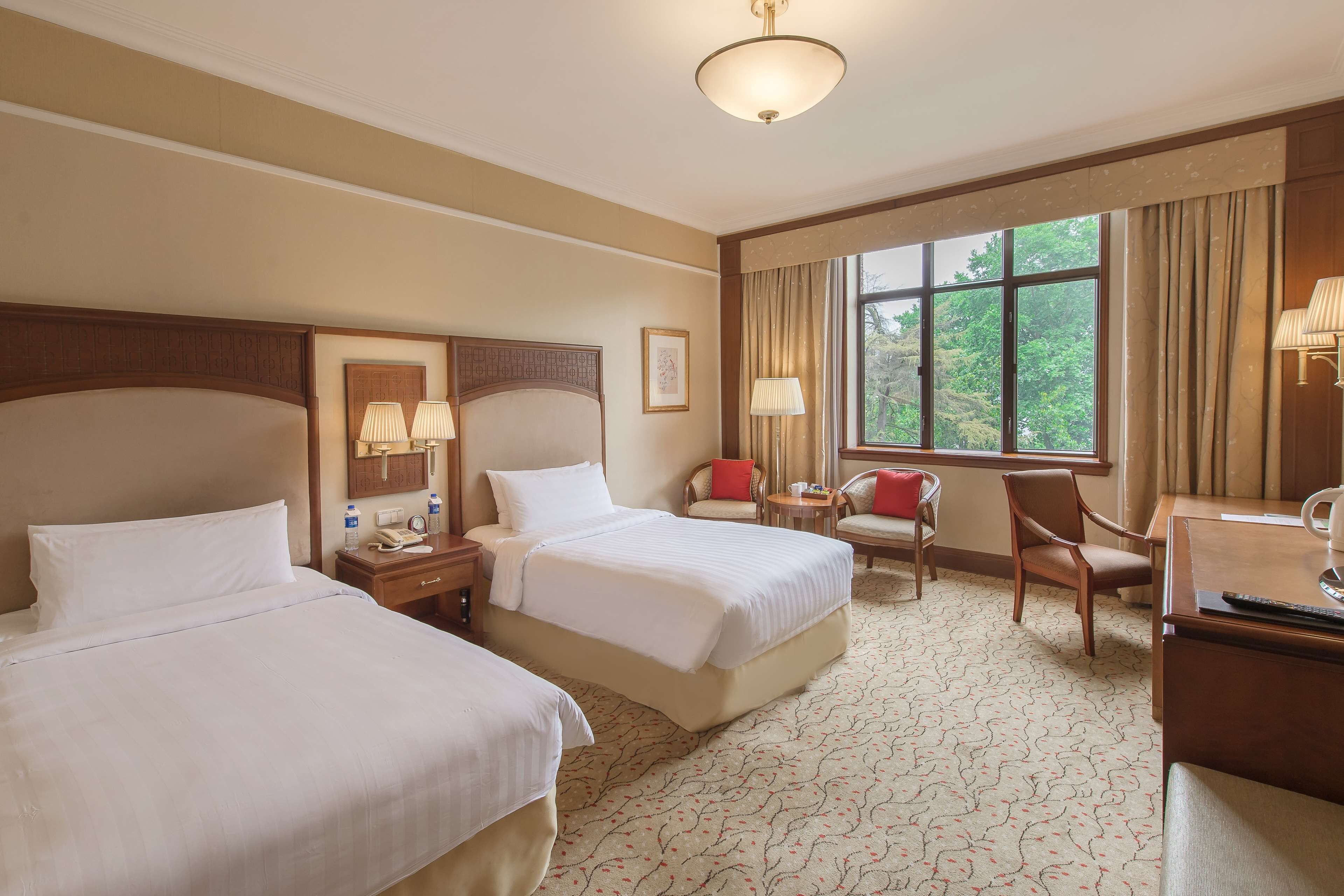 A hotel room with two beds, a desk, a chair, a window with curtains, and a ceiling light.