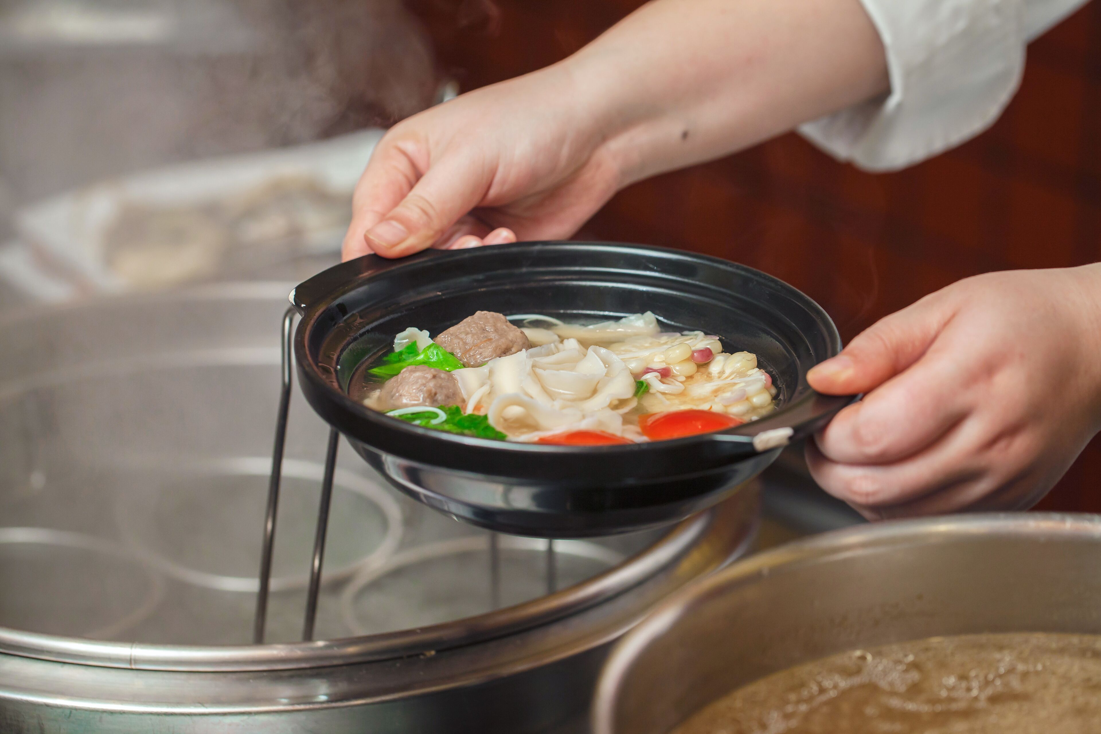 A person holding a bowl of soup with noodles and vegetables over a pot.