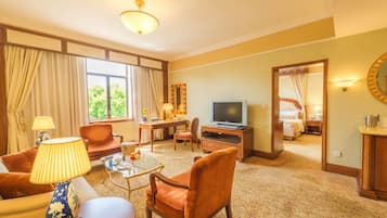 A hotel room with a sofa, armchair, TV, and a desk with a lamp.