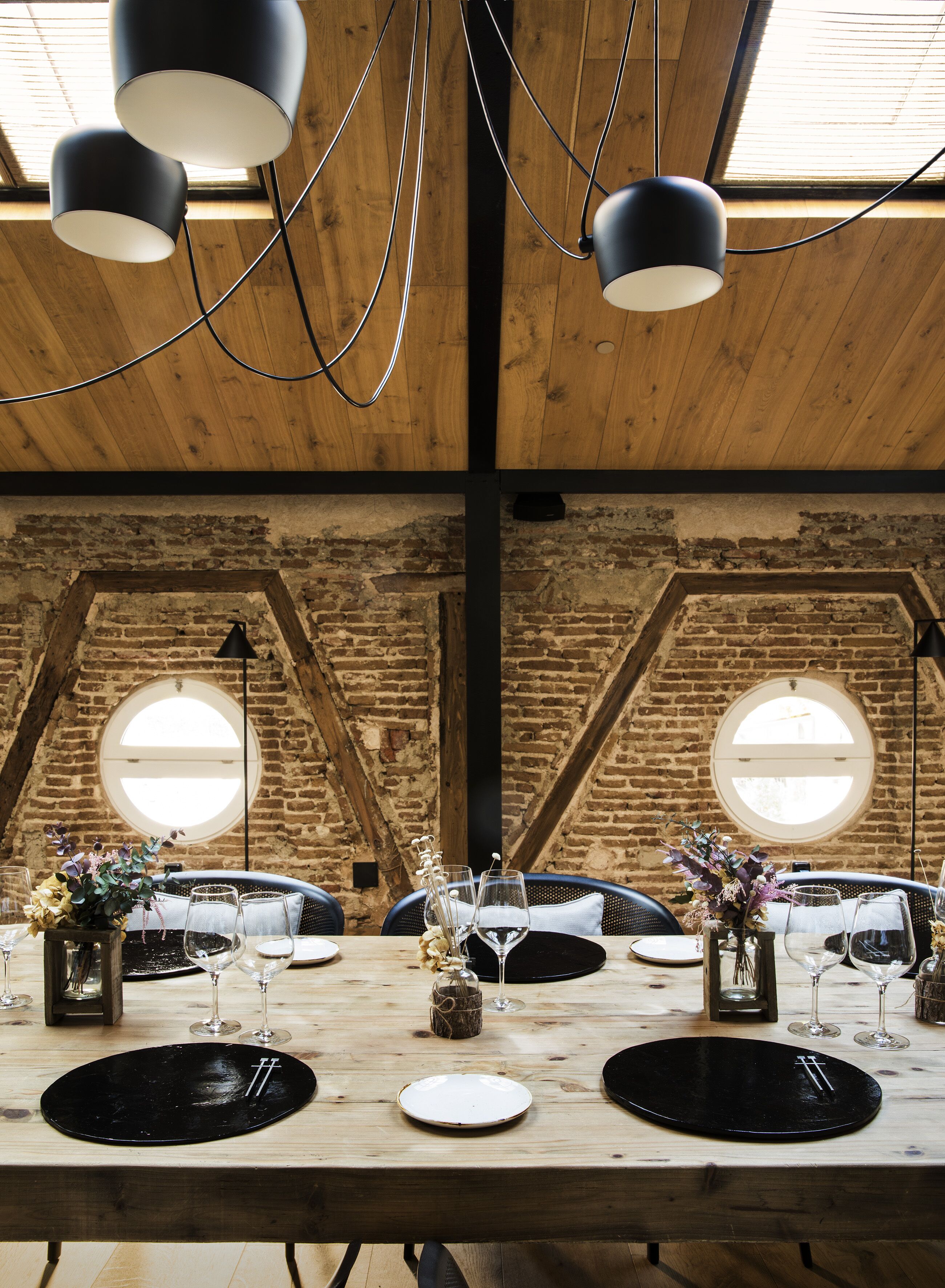 A dining area with a wooden table set for a meal, featuring black plates, wine glasses, and a centerpiece.
