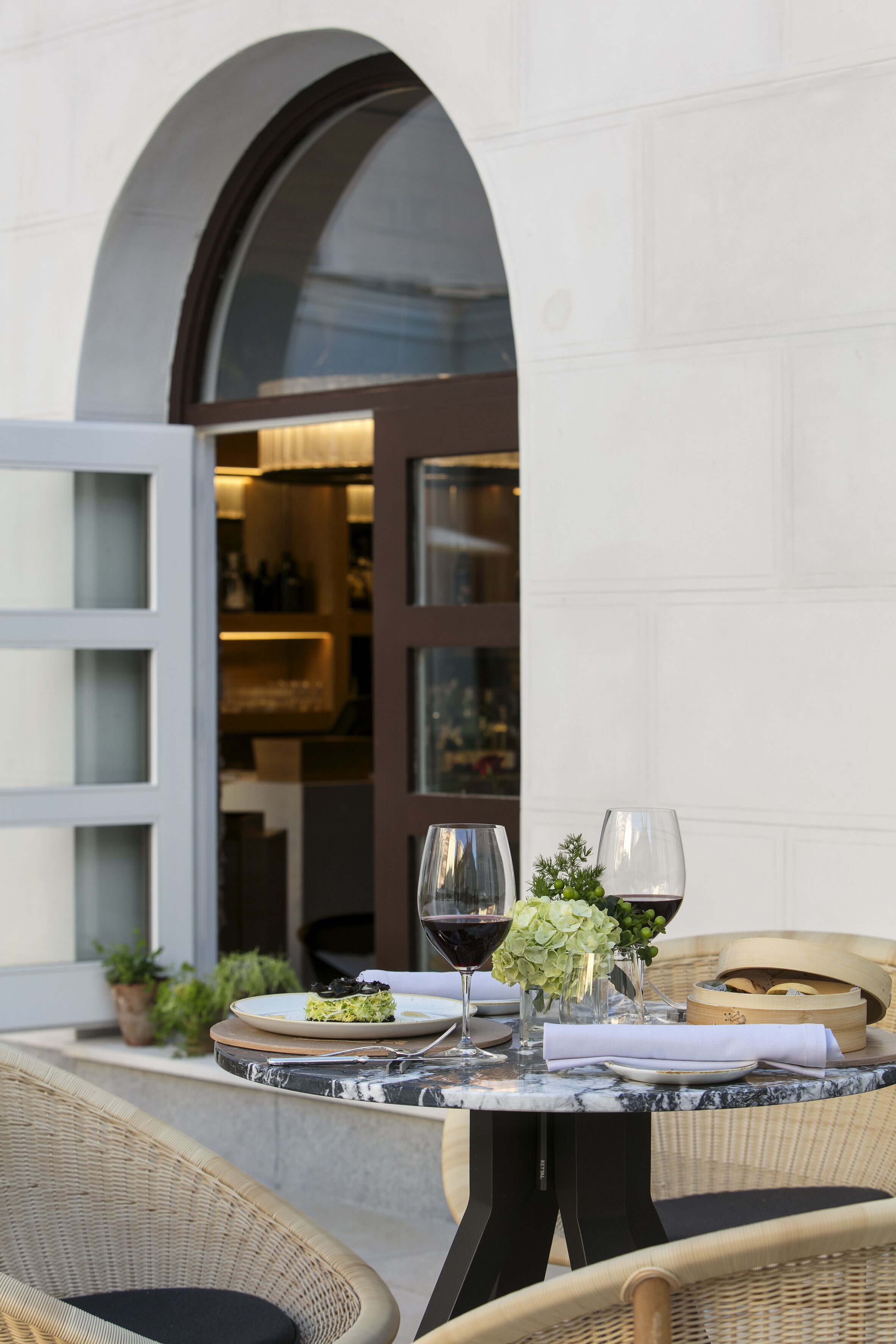 Un área de comedor al aire libre con sillas de mimbre, una mesa de mármol y una copa de vino tinto.