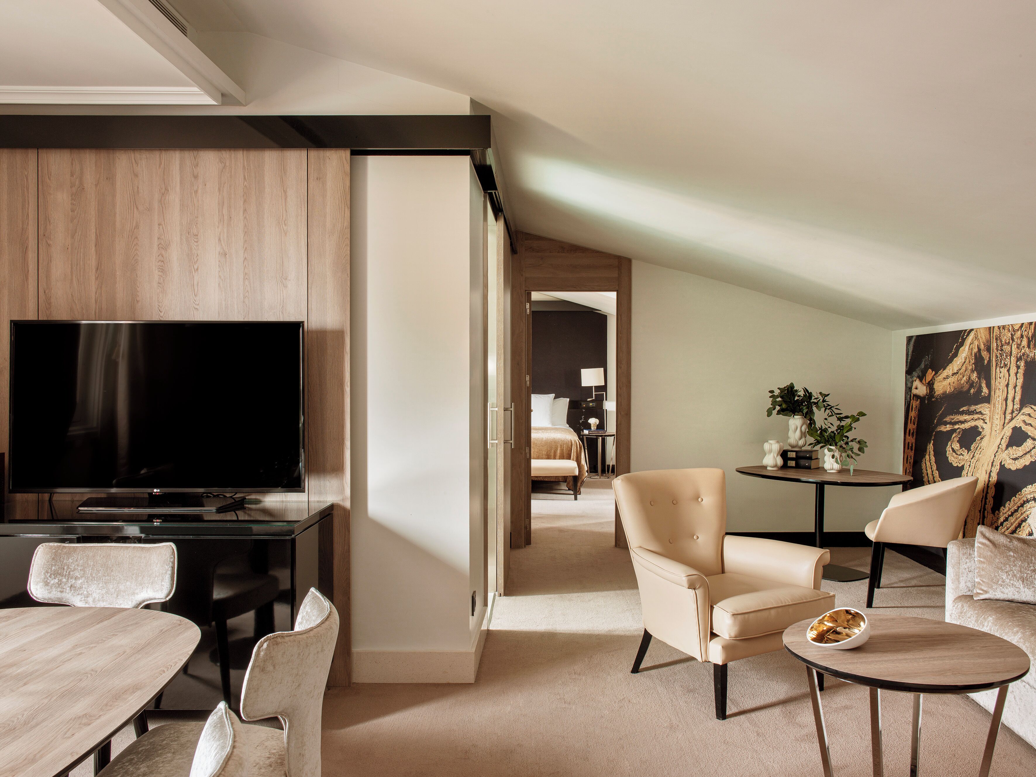 A modern hotel room with a flat-screen TV, a dining area with a table and chairs, a sofa, and a small side table with a bowl.