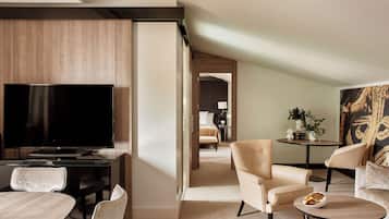 A modern hotel room with a flat-screen TV, a dining area with a table and chairs, a sofa, and a small side table with a bowl.