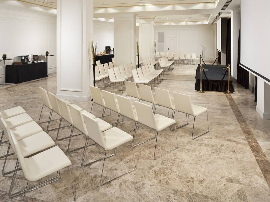 Un salón de conferencias con filas de sillas blancas, una mesa negra con un micrófono y una pantalla al frente.