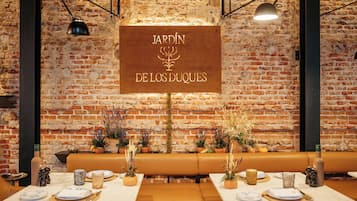 A restaurant interior with a brick wall backdrop, a sign reading "JARDÍN DE LOS DUQUES", and a dining area set with tables, chairs, and potted plants.