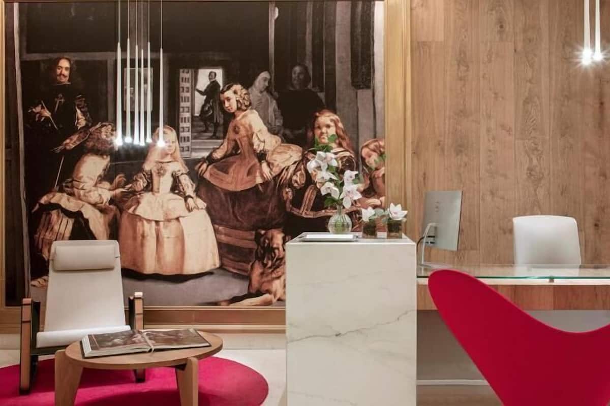 A modern interior with a large mirror reflecting a classical painting, a marble reception desk, a red chair, and a wooden wall.