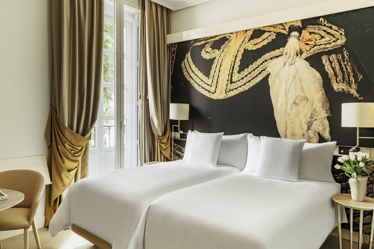 A hotel room with a large bed, a desk, a chair, a lamp, and a wall painting.