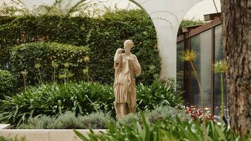 Una estatua en un jardín con abundante vegetación y un edificio moderno al fondo.