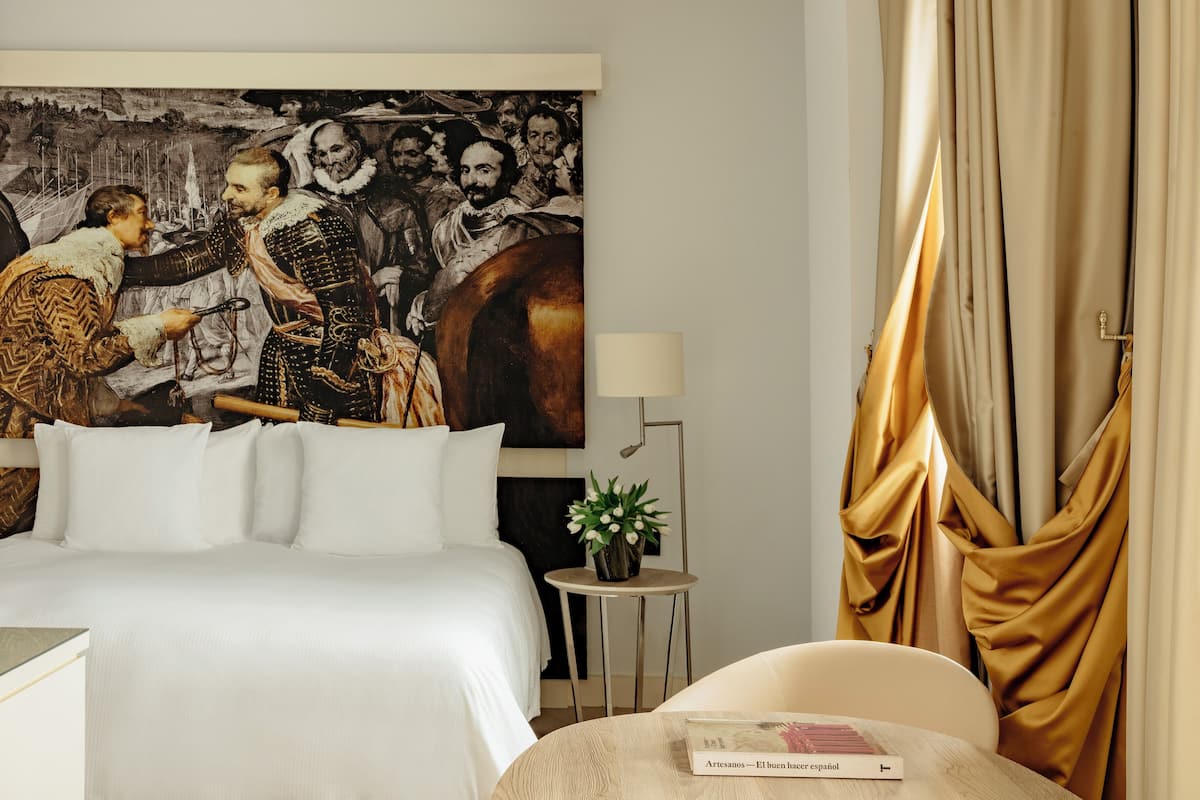 A hotel room with a large bed, a small bedside table with a lamp, a chair, and a painting on the wall.