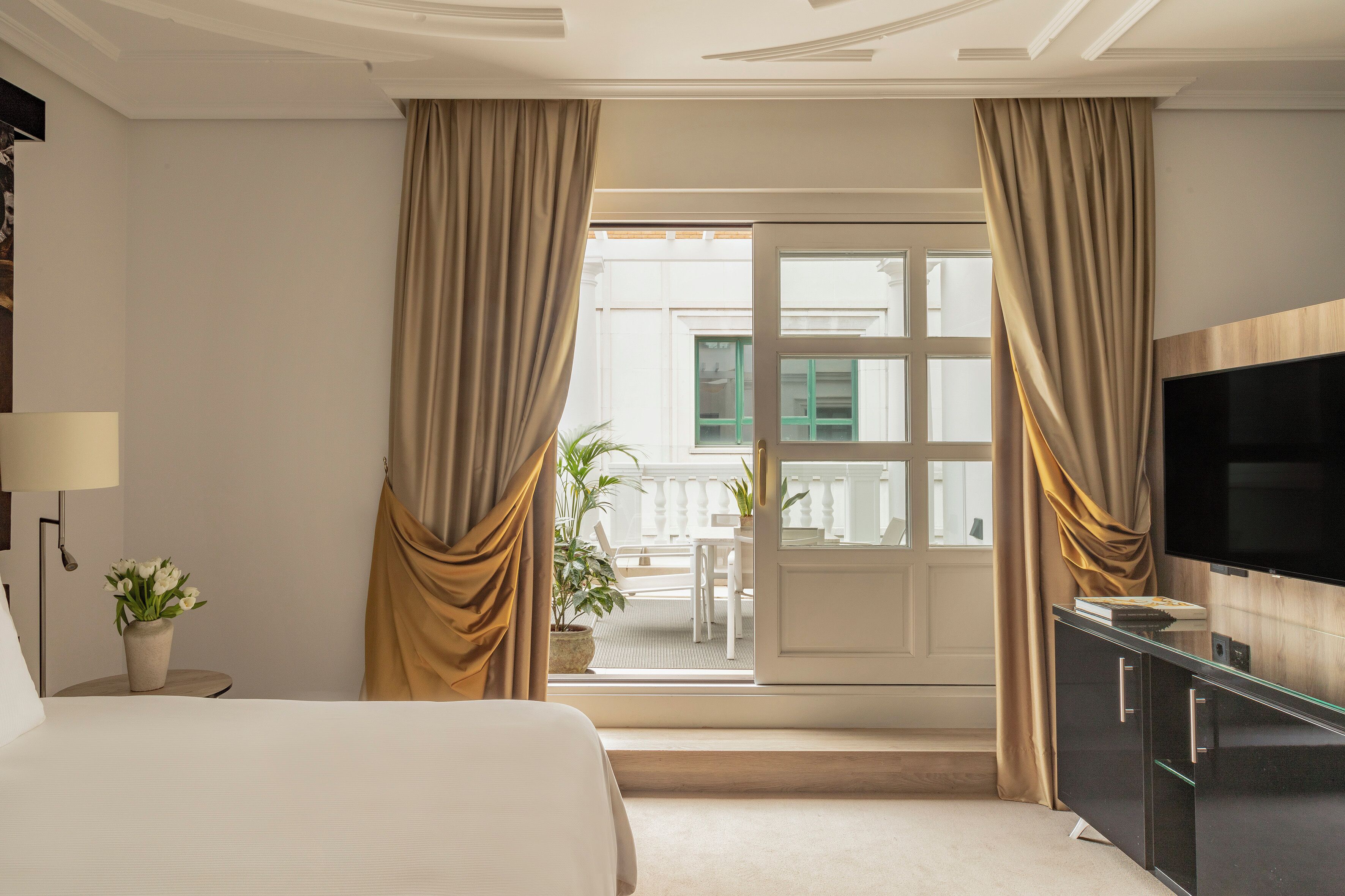 Habitación de hotel con cama, televisión, escritorio y balcón con cortinas.