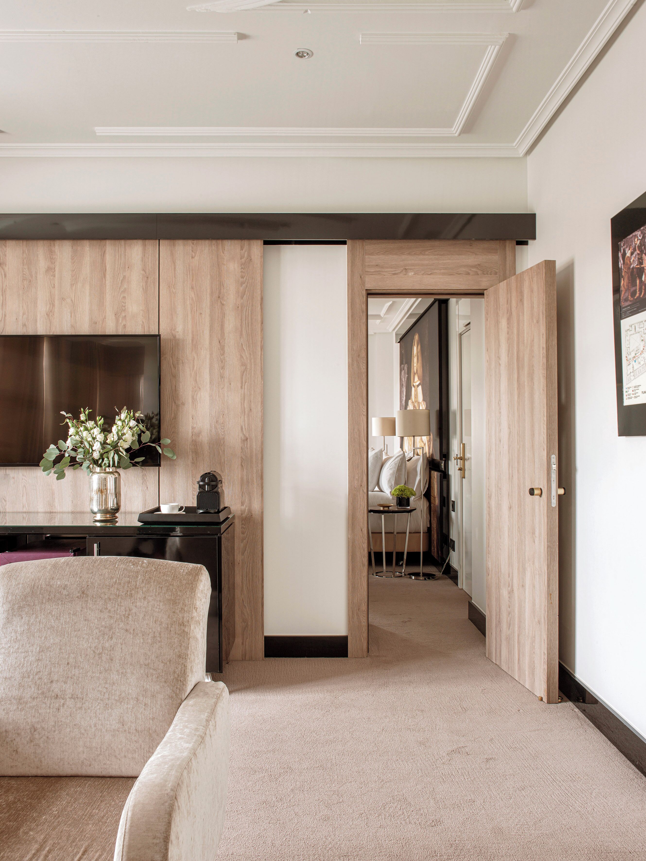 Una habitación de hotel moderna con un televisor de pantalla plana grande, un panel de madera decorativo, un sofá cómodo y una mesita de noche con un jarrón de flores.