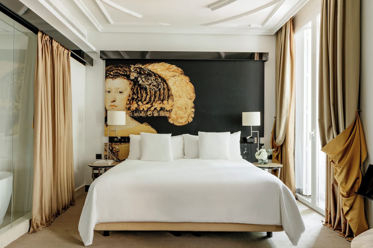 A large bed with white linens, a nightstand with a lamp, and a large painting on the wall.