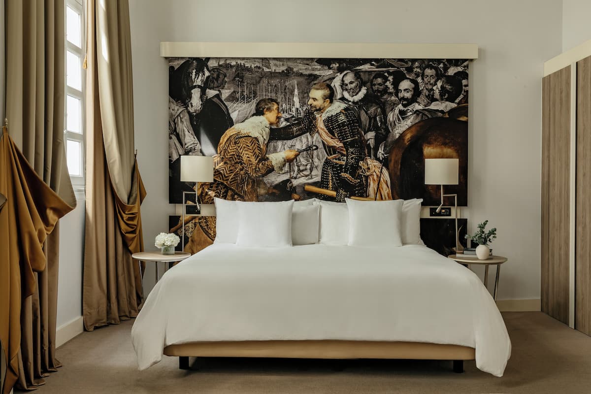 A large bed with white linens, two bedside tables with lamps, and a large framed artwork on the wall.