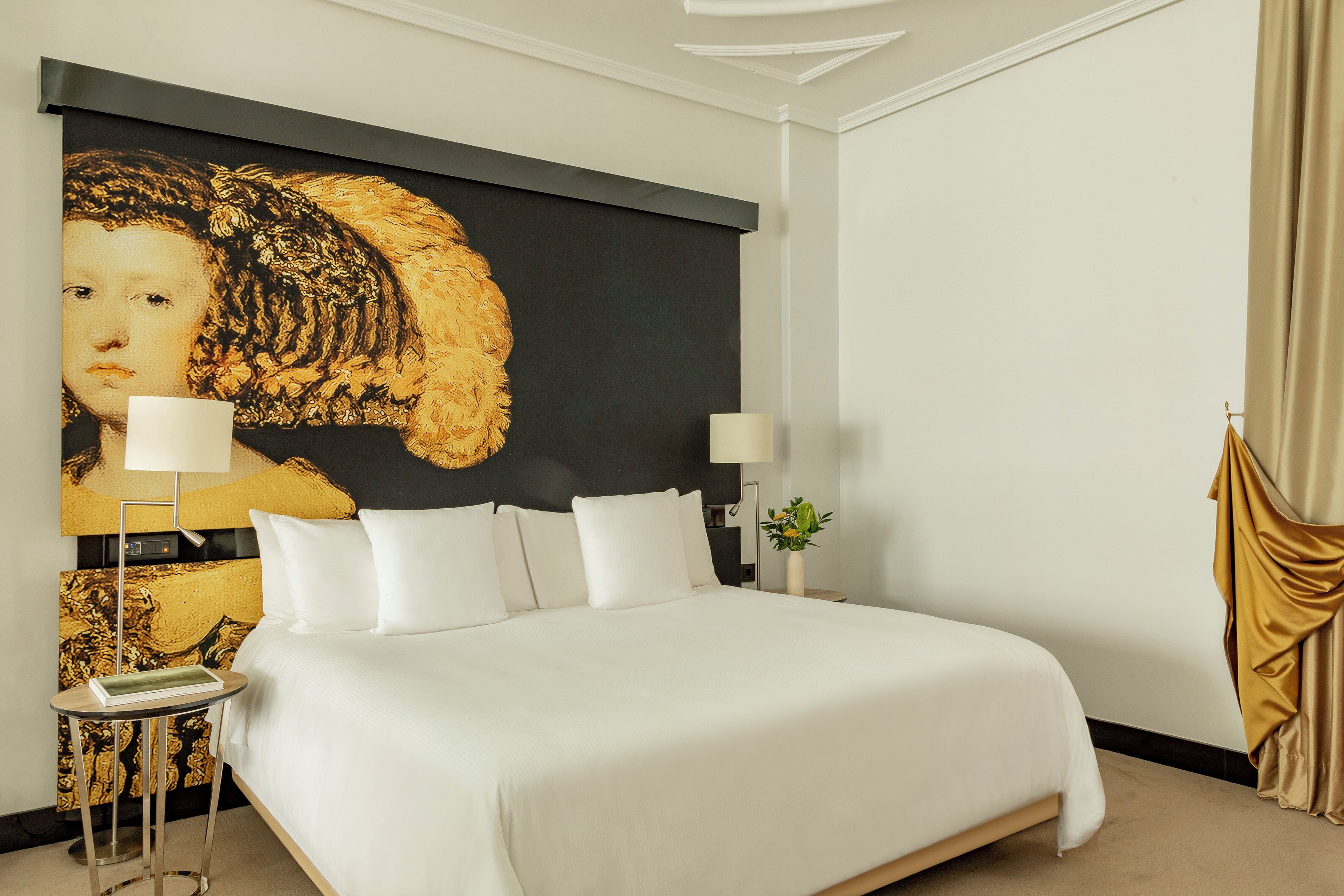 Un cuarto de hotel con una cama grande, dos lámparas de noche, una mesita auxiliar y un cuadro grande en la pared.