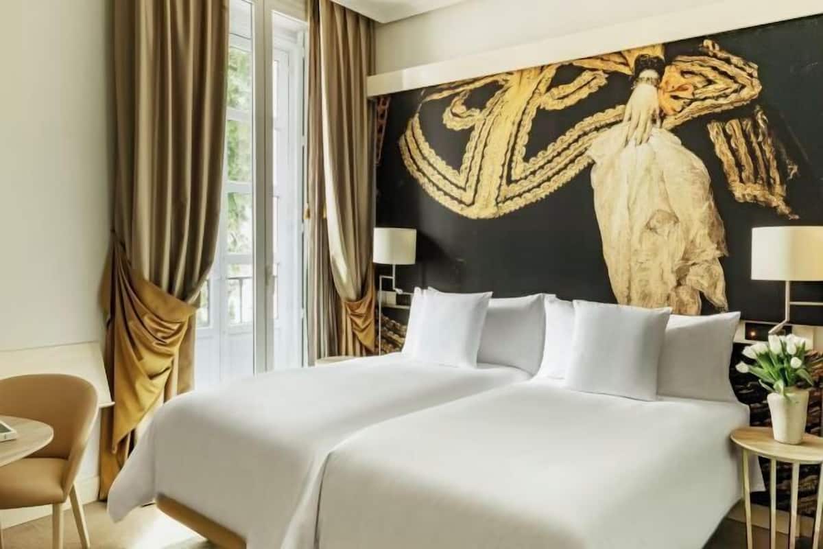 A hotel room with a large bed, a desk, a chair, a window with curtains, and a wall painting.