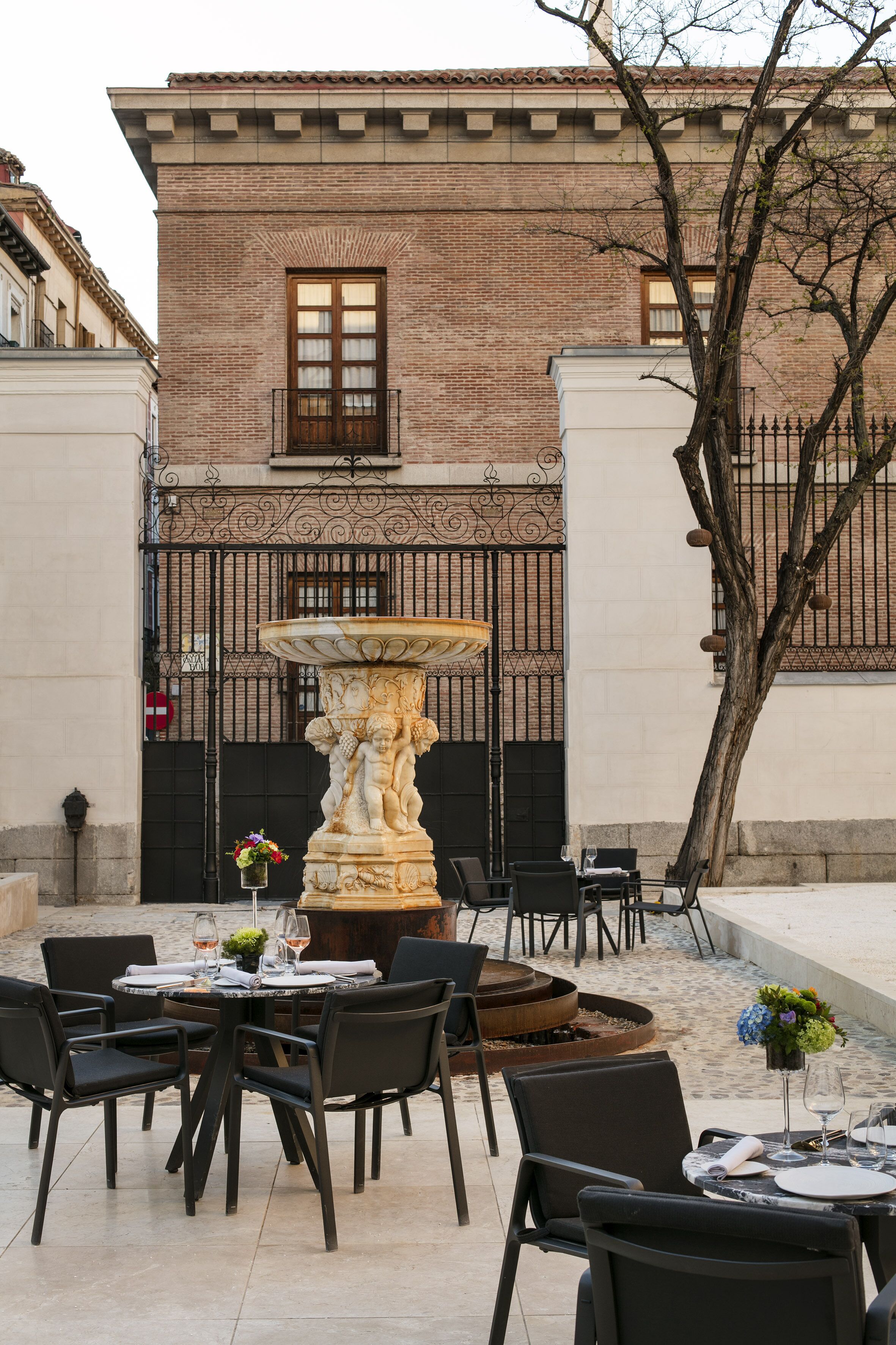 An outdoor dining area with a stone fountain, black chairs, and a brick building in the background.