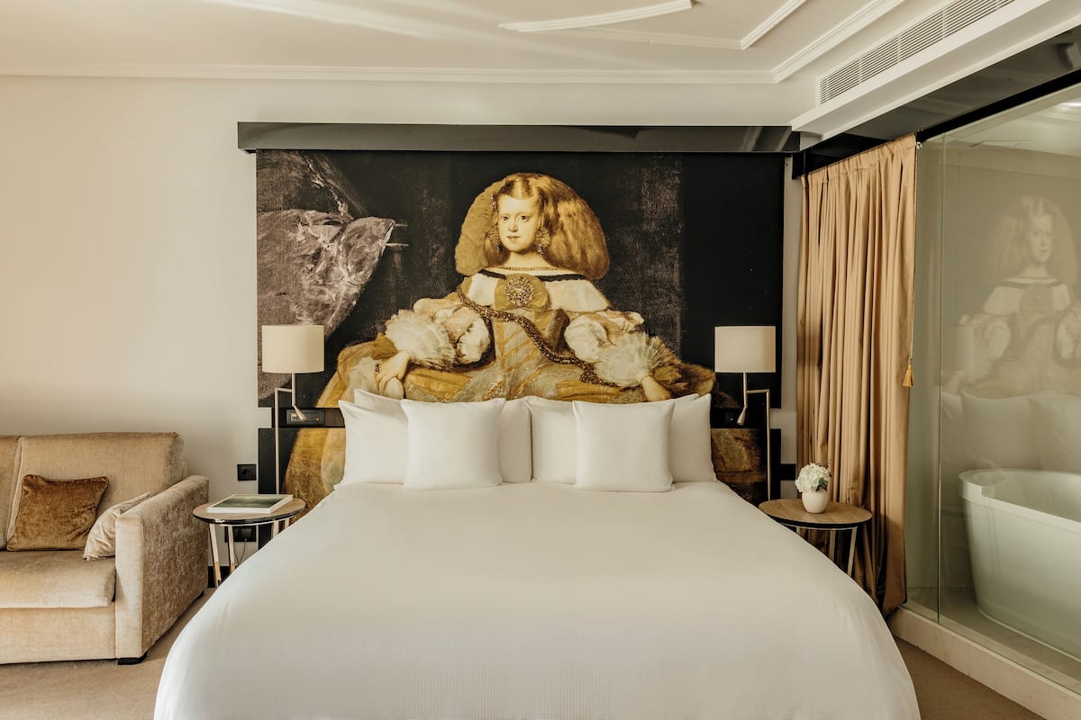 A large bed with white bedding, two bedside tables with lamps, a sofa, and a large painting on the wall.