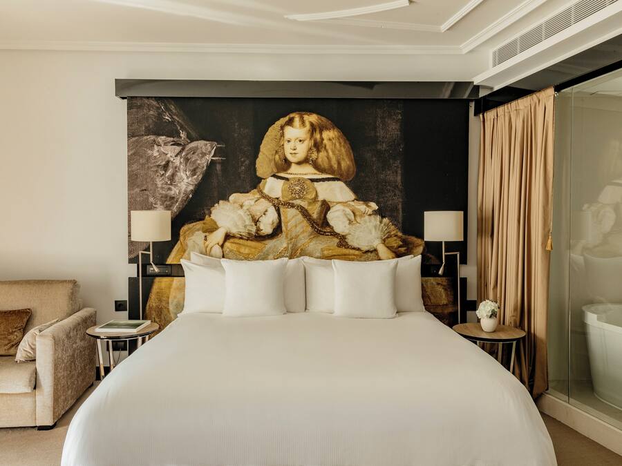 A large bed with white bedding, two bedside tables with lamps, a sofa, and a large painting on the wall.