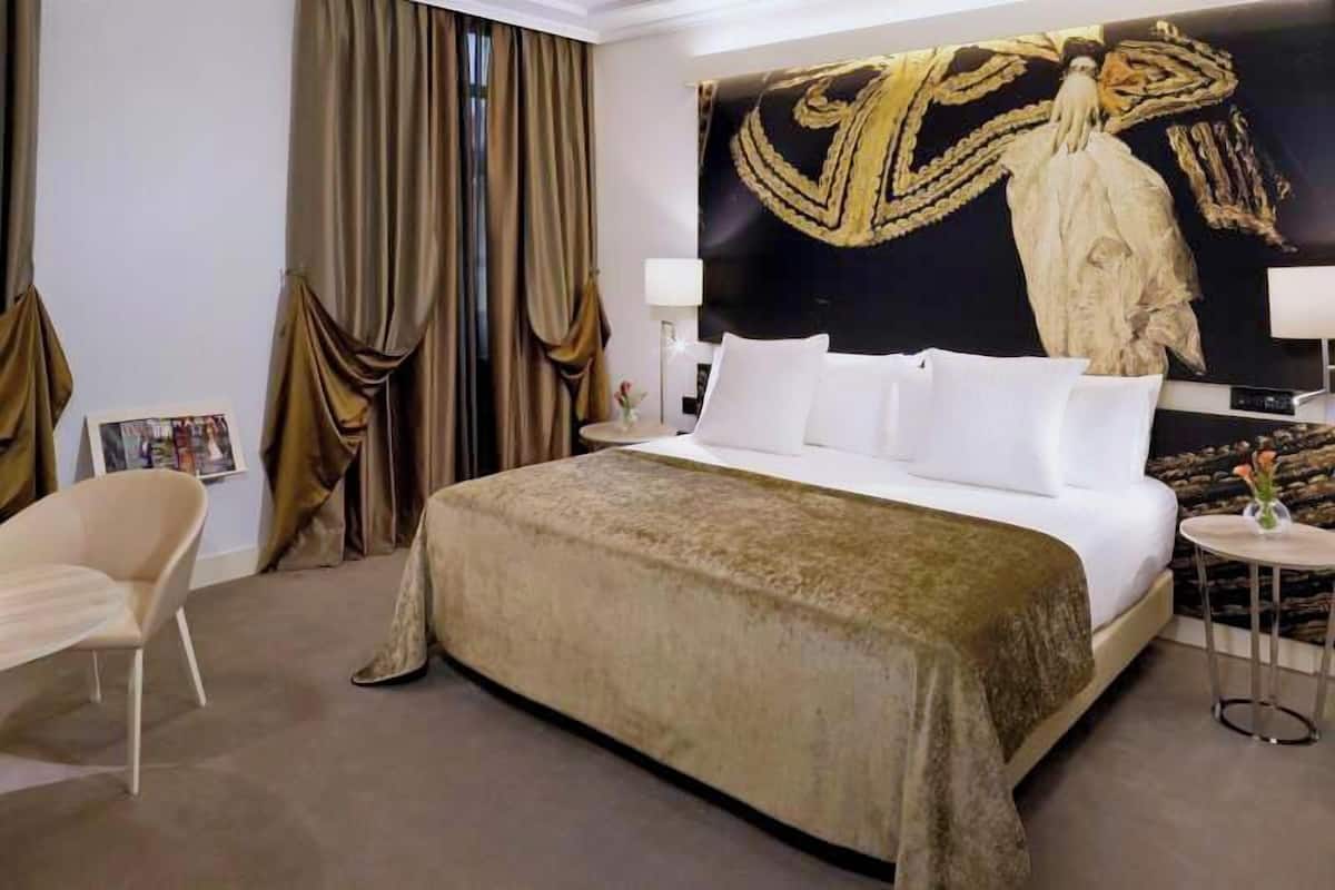 A hotel room with a large bed, a chair, a small table, and a wall with a large painting.