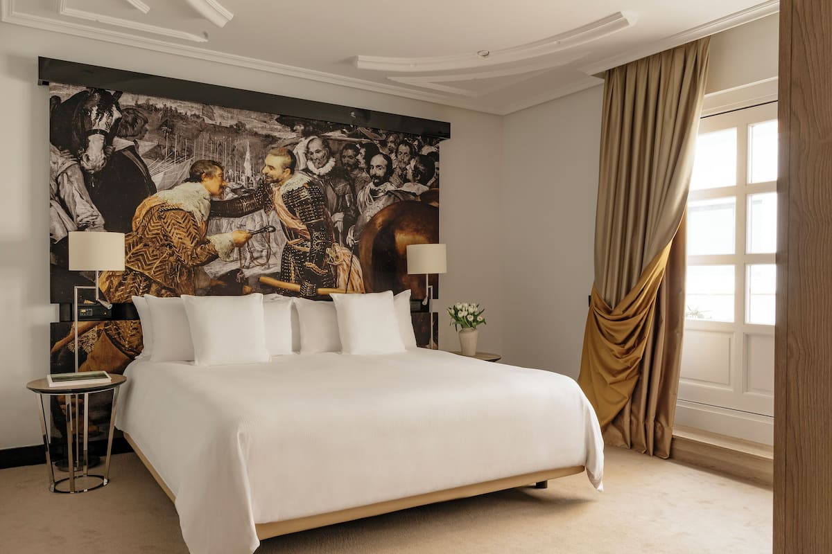 A hotel room with a large bed, a painting on the wall, a side table, and a window with curtains.