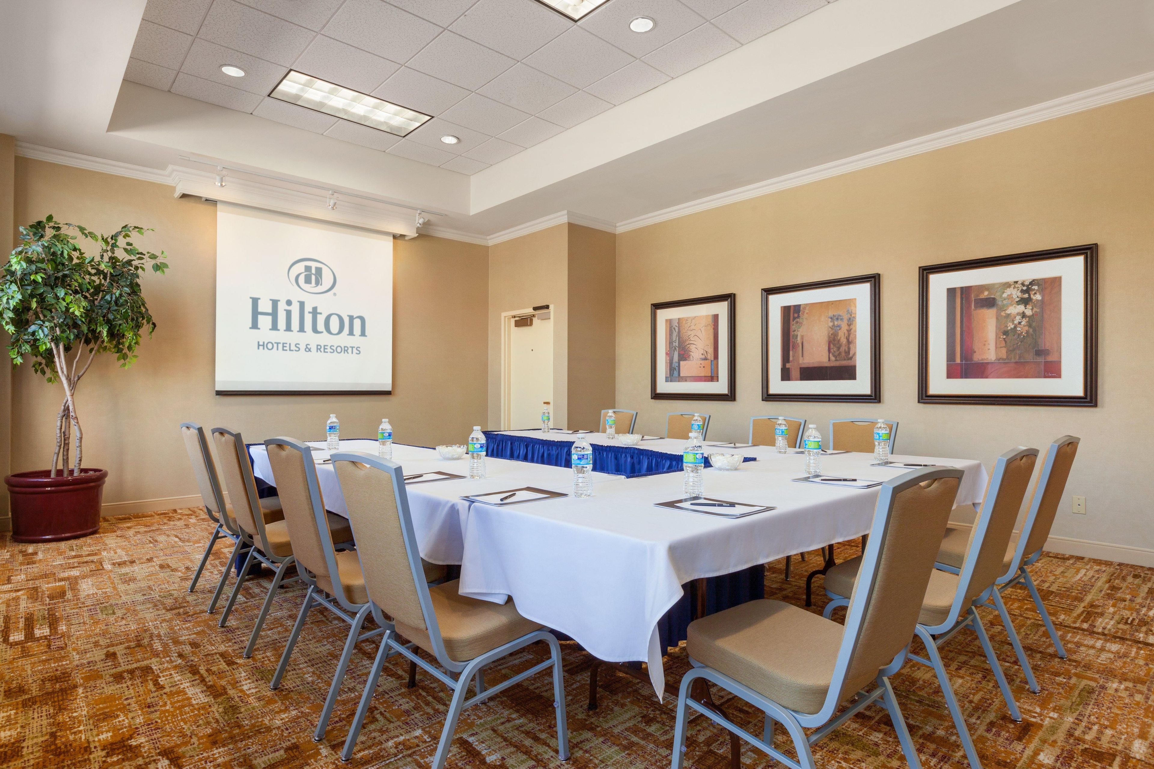 Une salle de conférence avec une longue table, des chaises, des bouteilles d’eau et un écran de projection affichant le logo de la chaîne Hilton Hotels & Resorts.