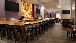 A modern bar area with a long counter, multiple bar stools, and a large wall-mounted TV. - Hilton Fort Wayne at the Grand Wayne Convention Center (Fort Wayne)