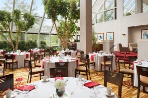 A spacious indoor dining area with multiple round tables, chairs, and a view of greenery through large windows.