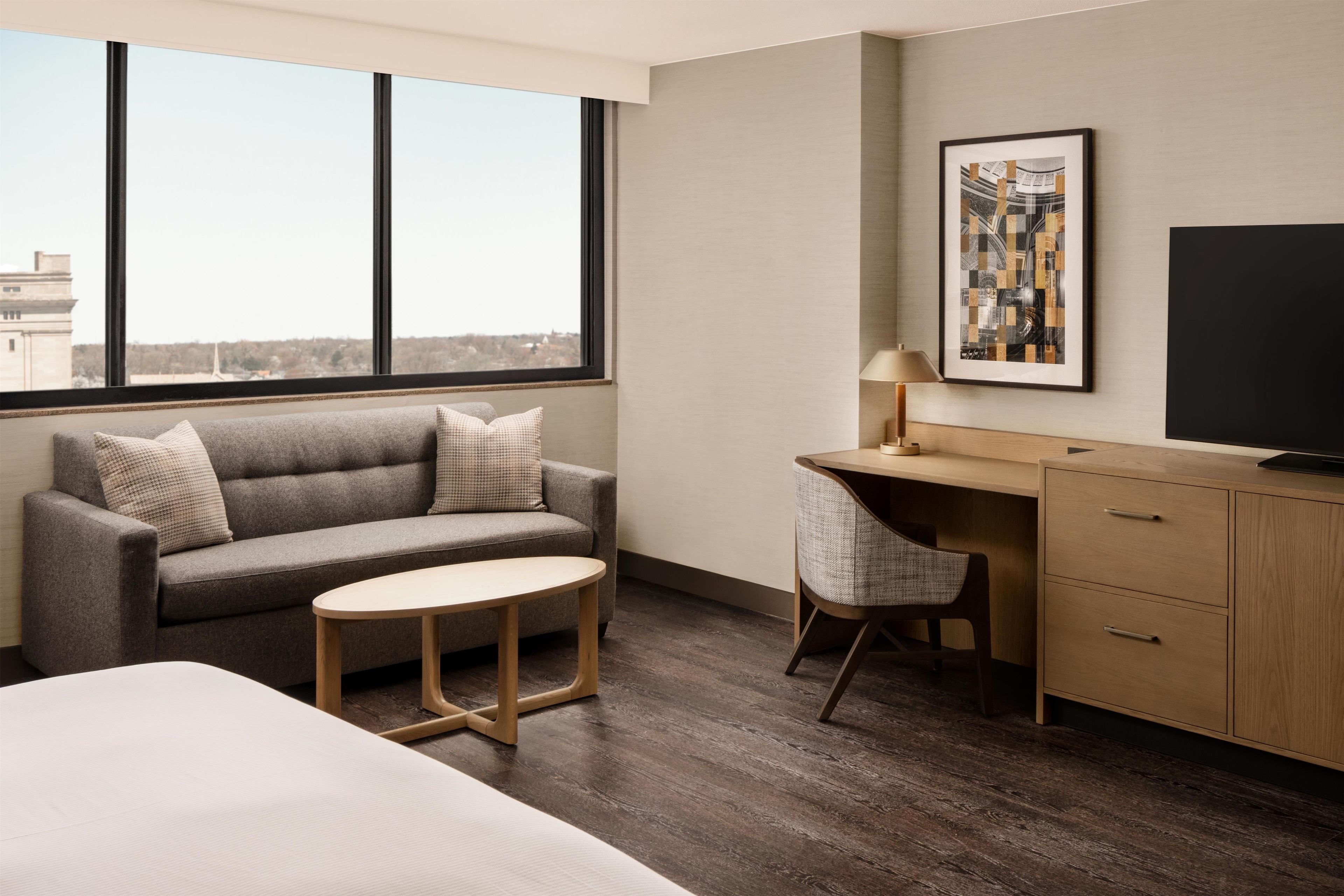 A hotel room with a sofa, a small round table, a desk with a chair, a television, and a large window offering a city view.