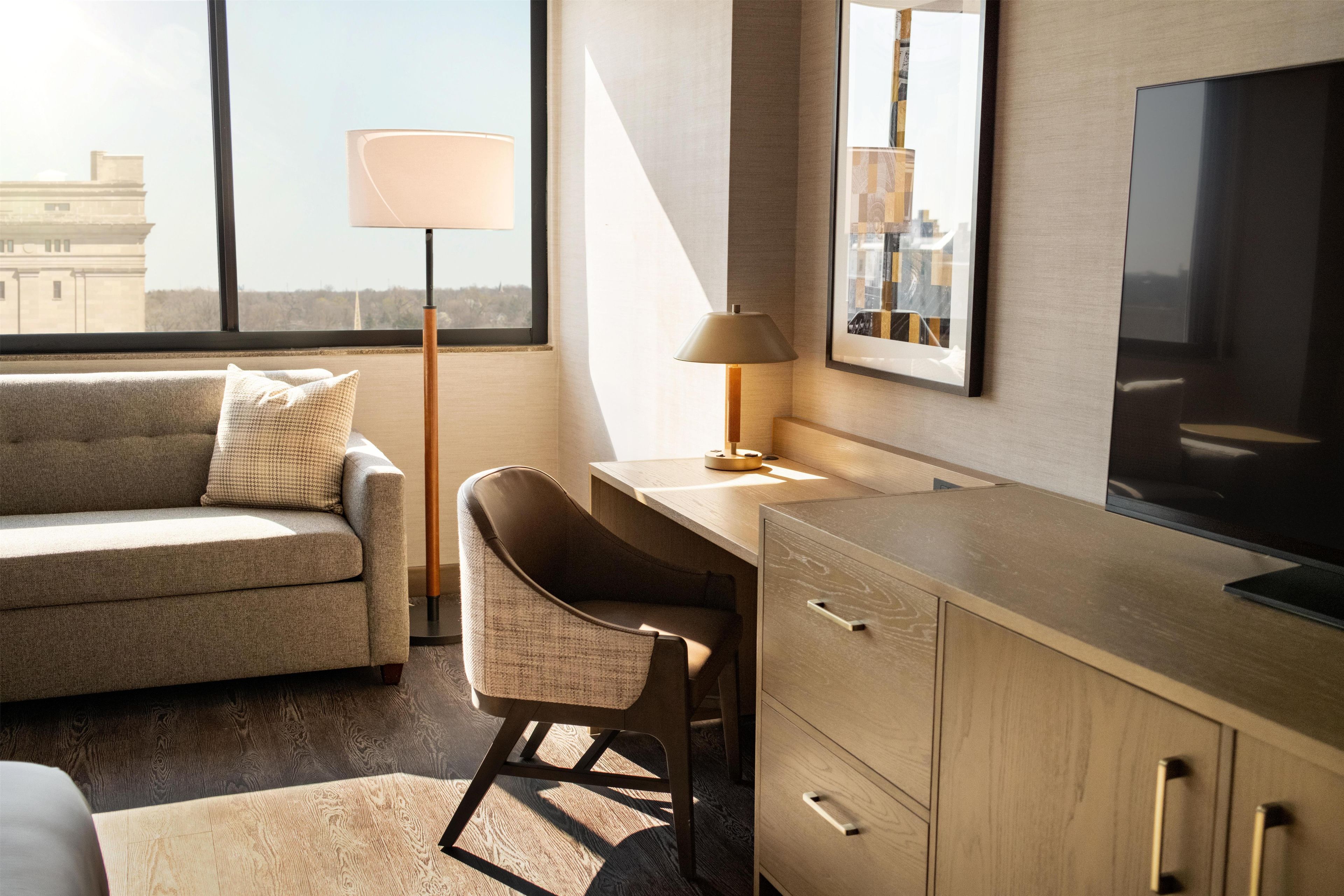 A modern hotel room with a sofa, a desk, a chair, a TV, and a large window.