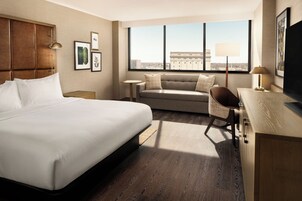 Habitación de hotel con una cama grande, un sofá, un escritorio y una televisión. Hay cuadros en la pared y una ventana con vistas a un edificio.