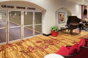 A hotel lobby with a grand piano, red chairs, a potted plant, and a carpet with a red and gold pattern.