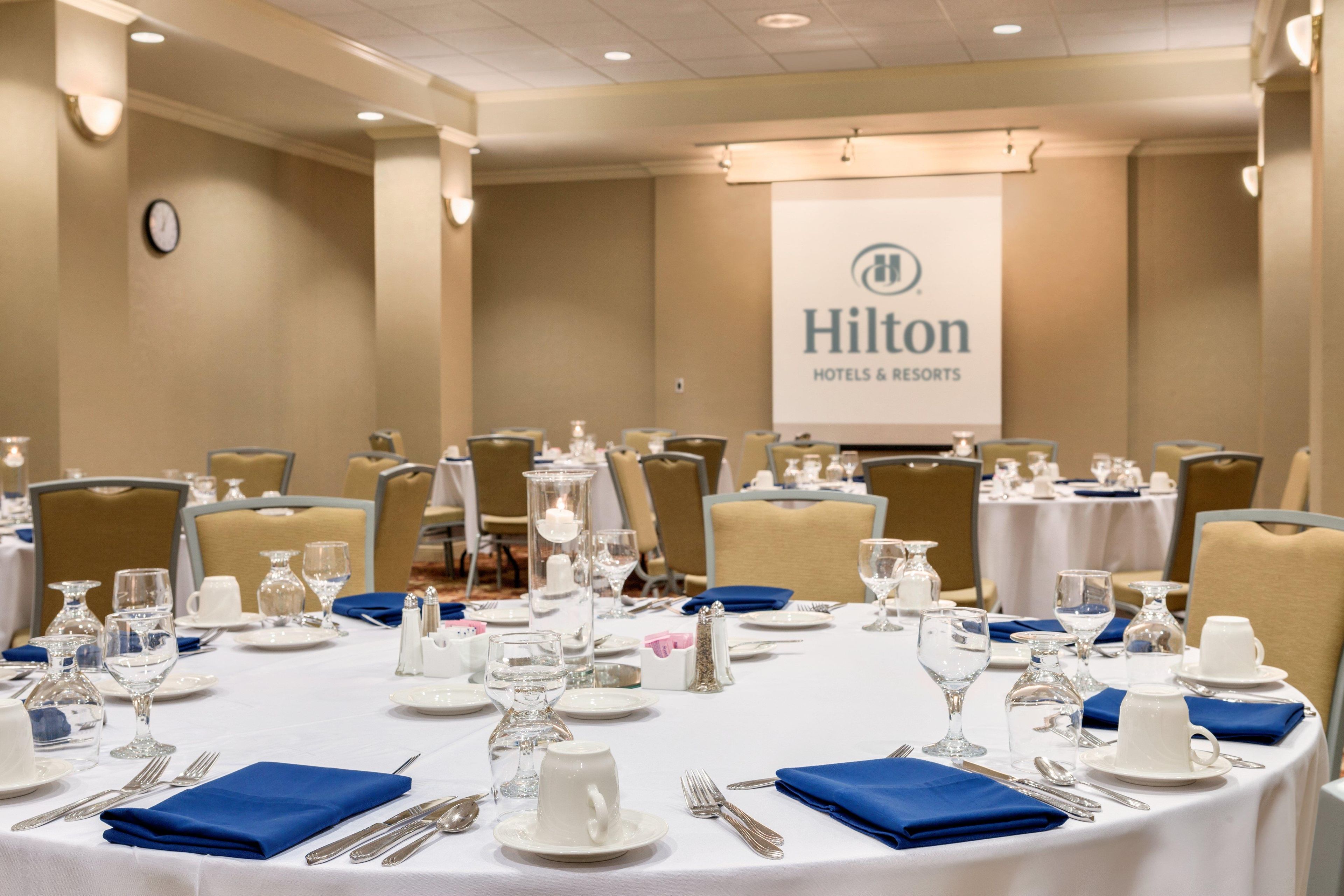 A conference room with round tables set for a meeting, equipped with nameplates, water glasses, and a projector screen displaying the Hilton Hotels & Resorts logo.