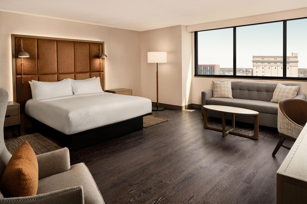 A hotel room with a large bed, a sofa, a small table, and a view of a building through the window.