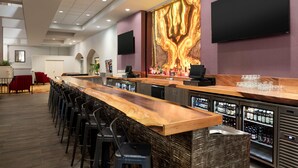 A bar area with a long wooden counter, multiple stools, and a wall-mounted TV. - Hilton Fort Wayne at the Grand Wayne Convention Center (Fort Wayne)