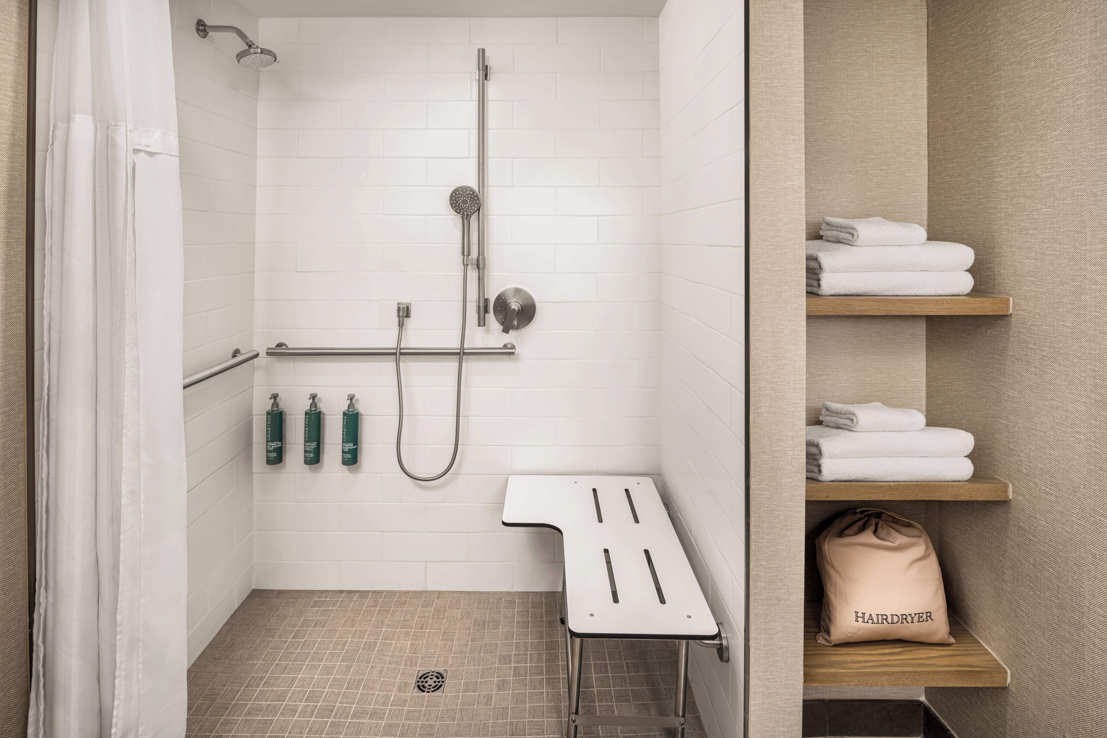 A shower area with a handheld showerhead, a wall-mounted showerhead, and a handheld shower attachment. There are three bottles of shampoo and conditioner on the wall. A white towel rack with folded towels is visible.