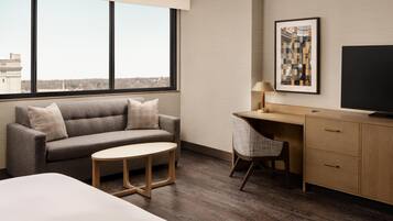 A hotel room with a sofa, a small round table, a desk with a chair, a television, and a large window offering a city view.