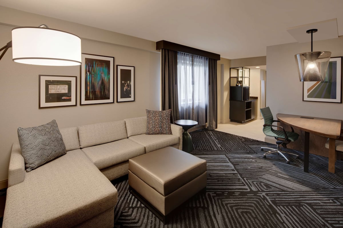A hotel room with a sectional sofa, a small table, a desk, and a chair. There are framed pictures on the wall and a large window with curtains.