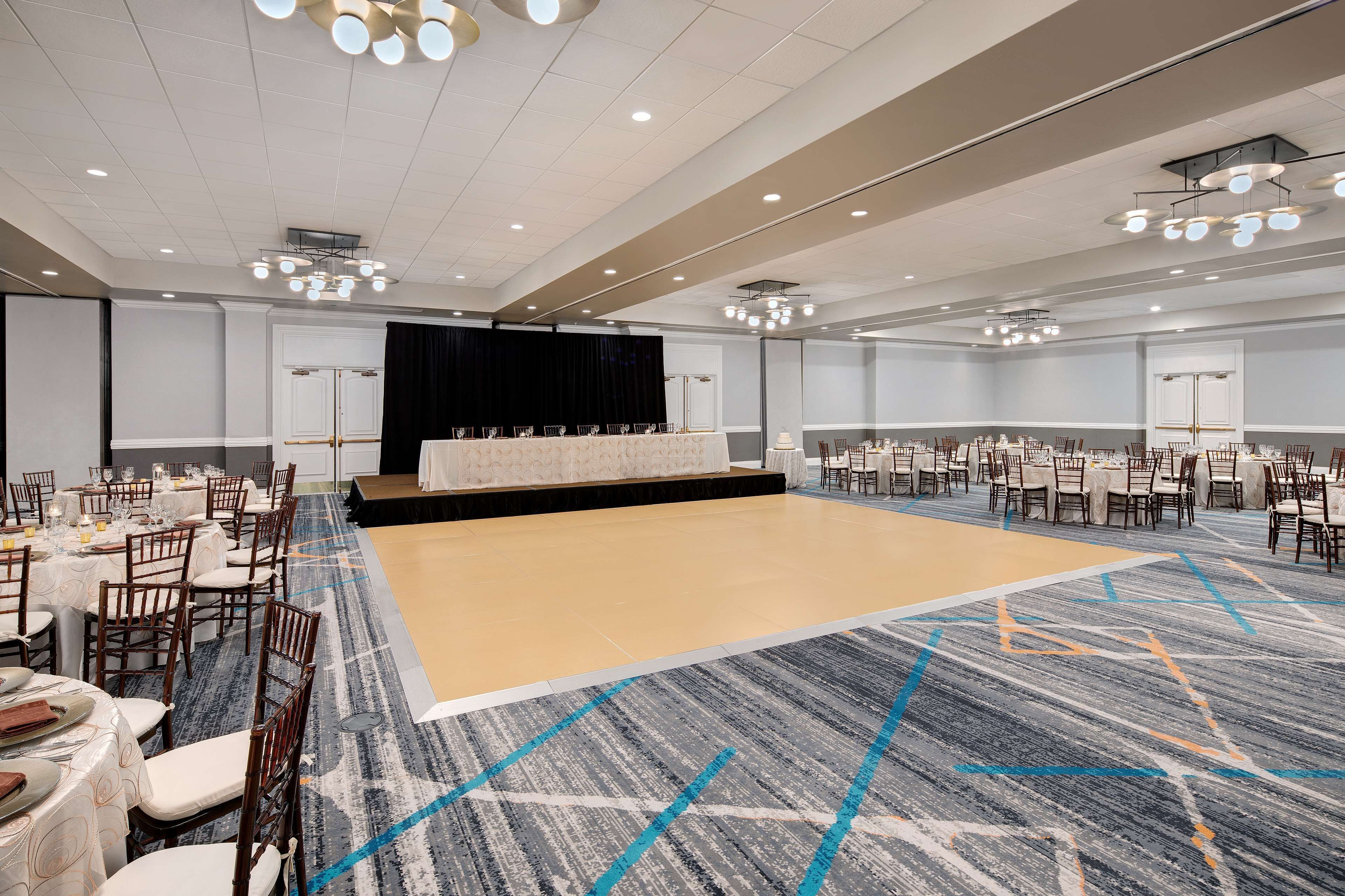A large conference room with a stage, multiple tables with chairs, and a carpeted floor.