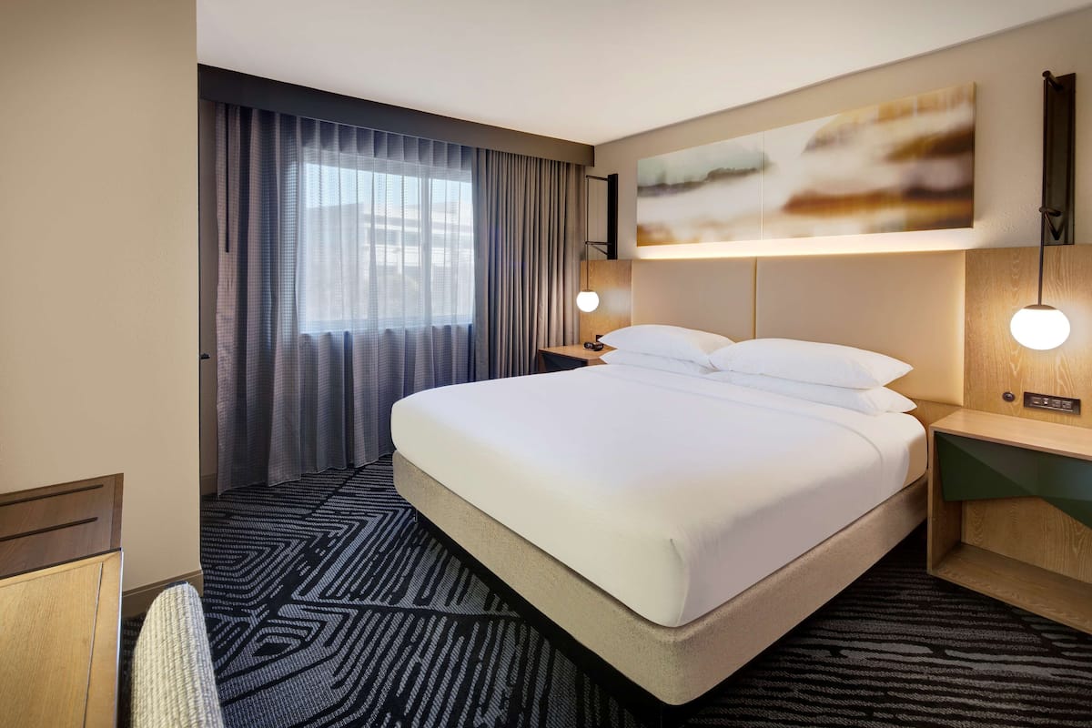 A hotel room with a large bed, a desk, a chair, a nightstand, a lamp, and a painting on the wall.