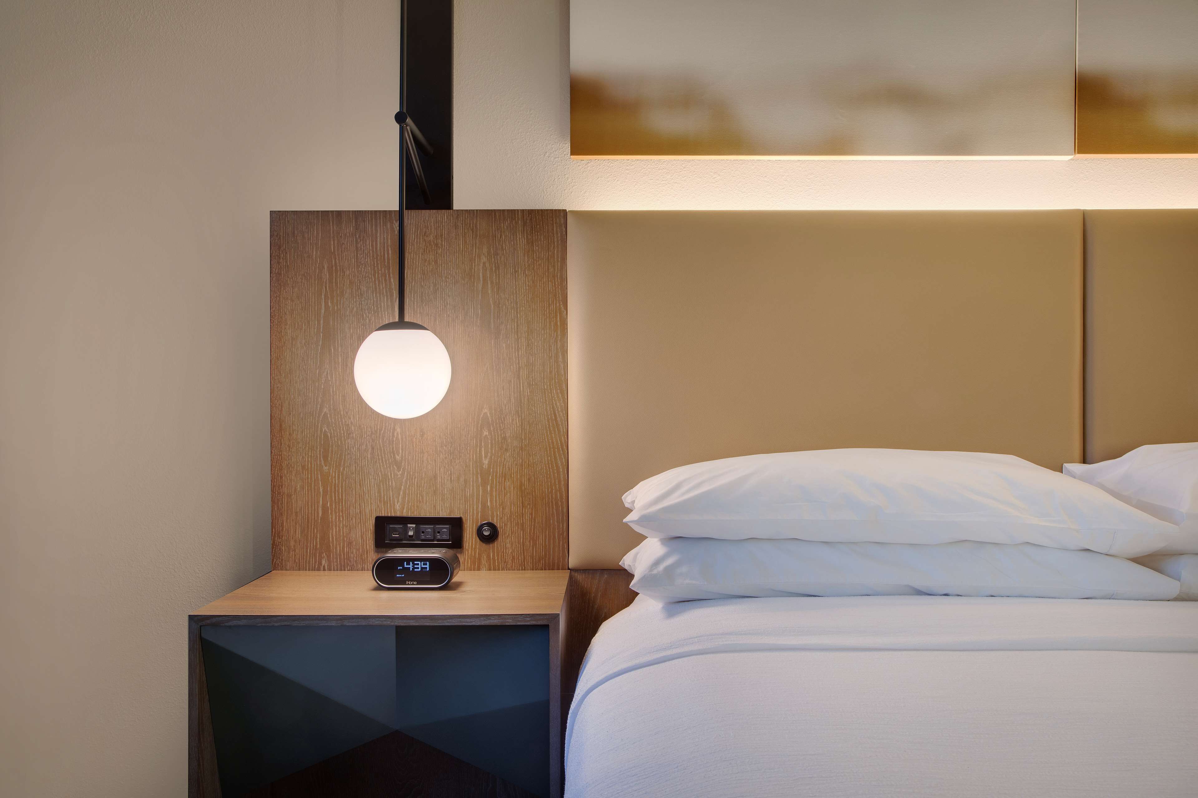 En pænt redt seng med hvide sengetæpper, et natbord med et ur og en lampe monteret på væggen.