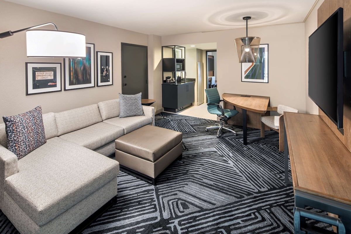 A modern hotel room with a sectional sofa, a small ottoman, a desk with a chair, a flat-screen TV, and a patterned carpet.