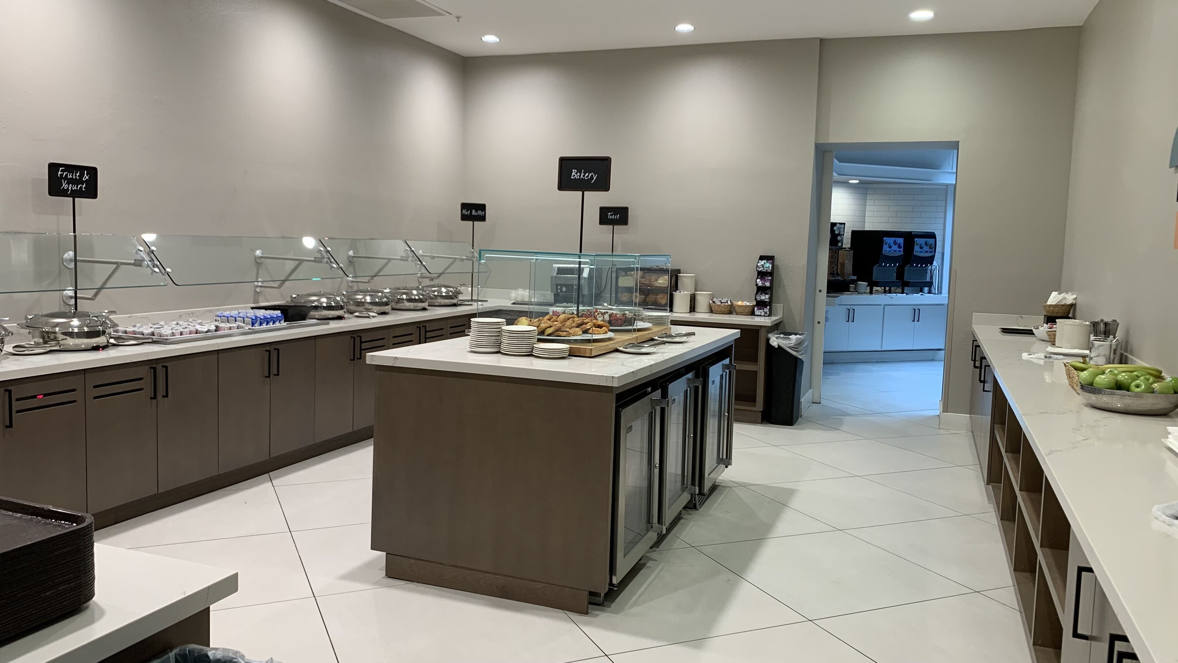 Een moderne keuken met een centraal eiland, diverse food stations aangeduid als 'Fruit & Yogurt', 'Warm Buffet' en 'Bakkerij', en uitzicht op de eetruimte.