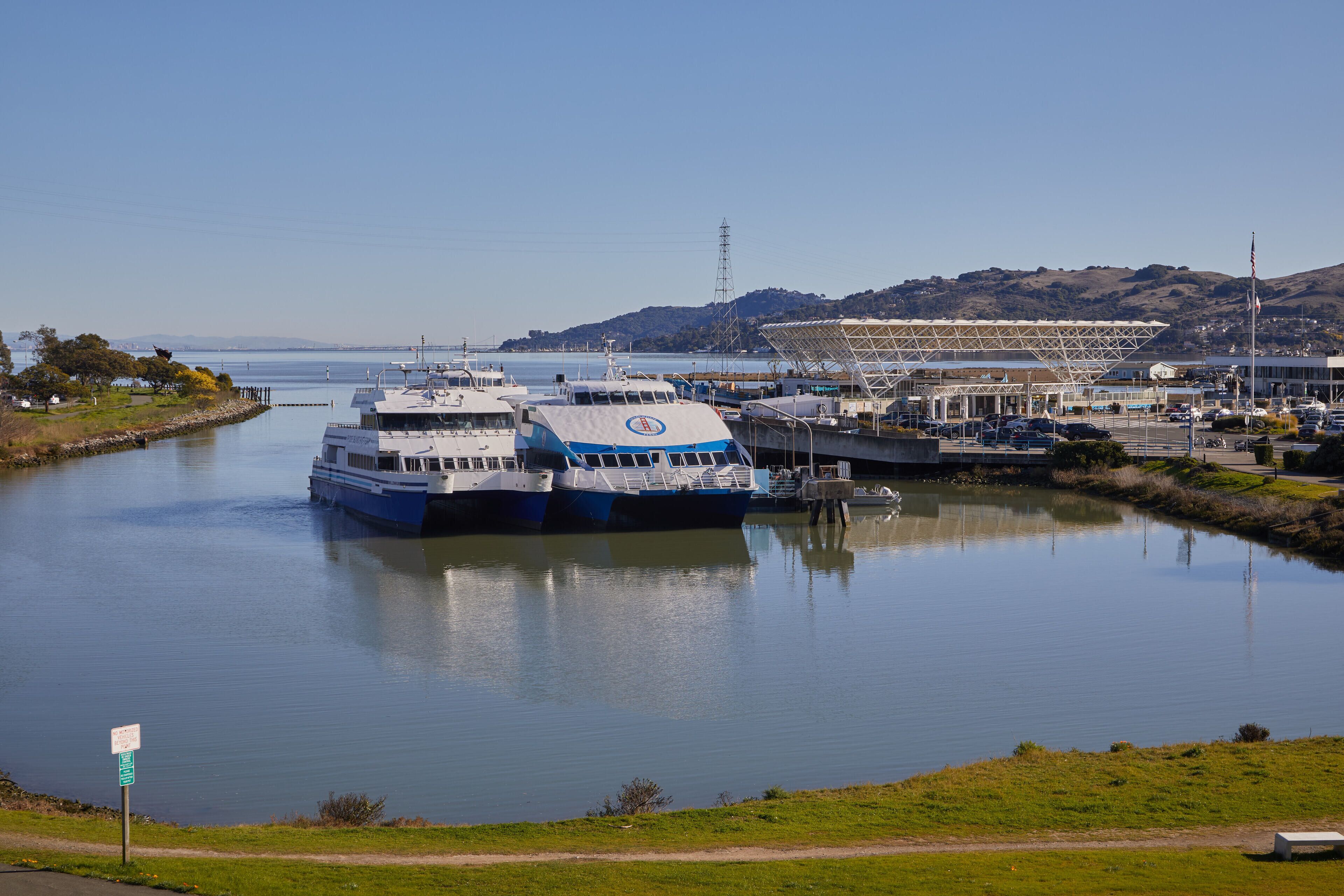 A marina with boats docked, a large building with a unique roof structure, and a grassy area in the foreground.