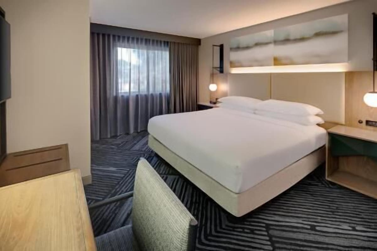 Habitación de hotel con una cama grande, un escritorio, una silla, un televisor, una ventana con cortinas y una lámpara fijada a la pared.