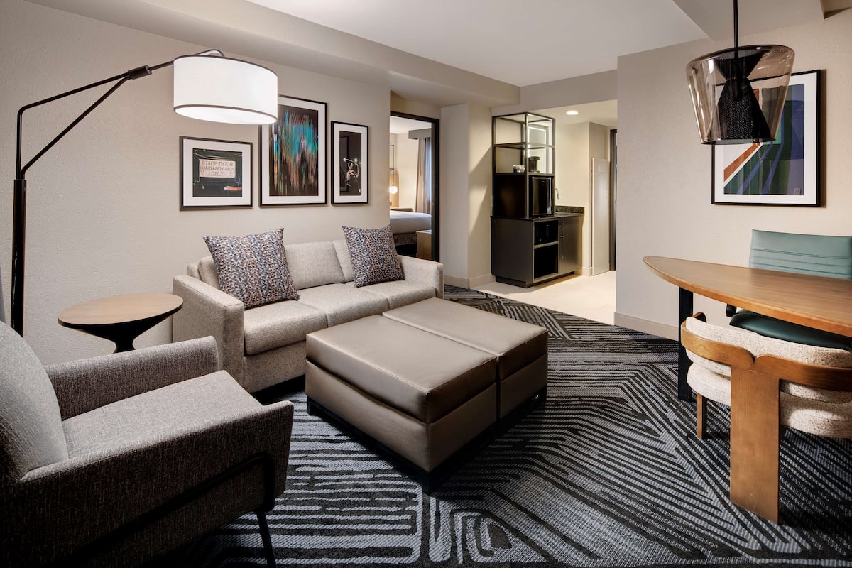 A modern hotel room with a sofa, ottoman, and a dining table. There is a lamp, framed artwork on the wall, and a TV in the background.